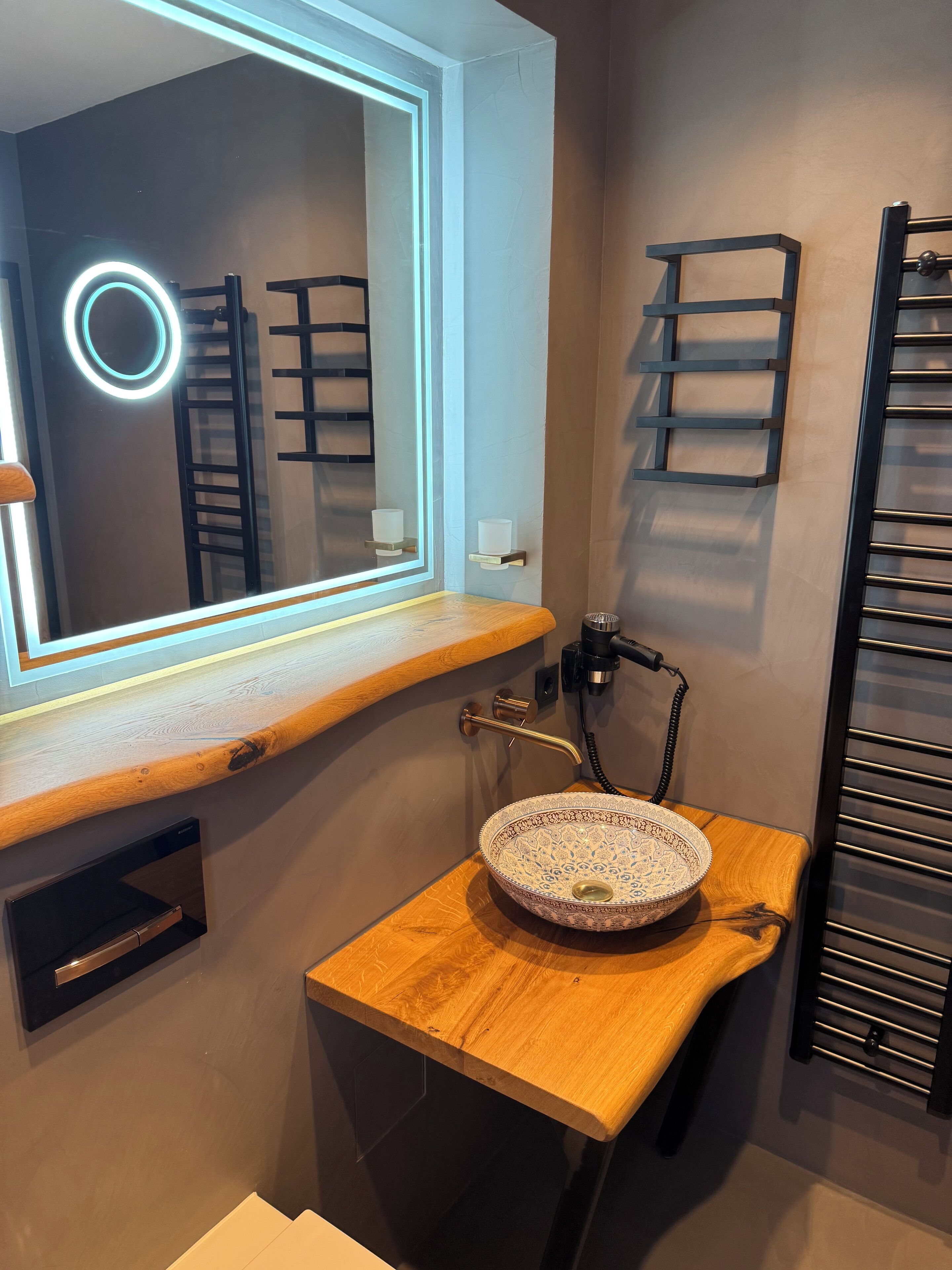 A bathroom with a wooden vanity, a round mirror with a light, a sink with a decorative bowl, and towel racks.