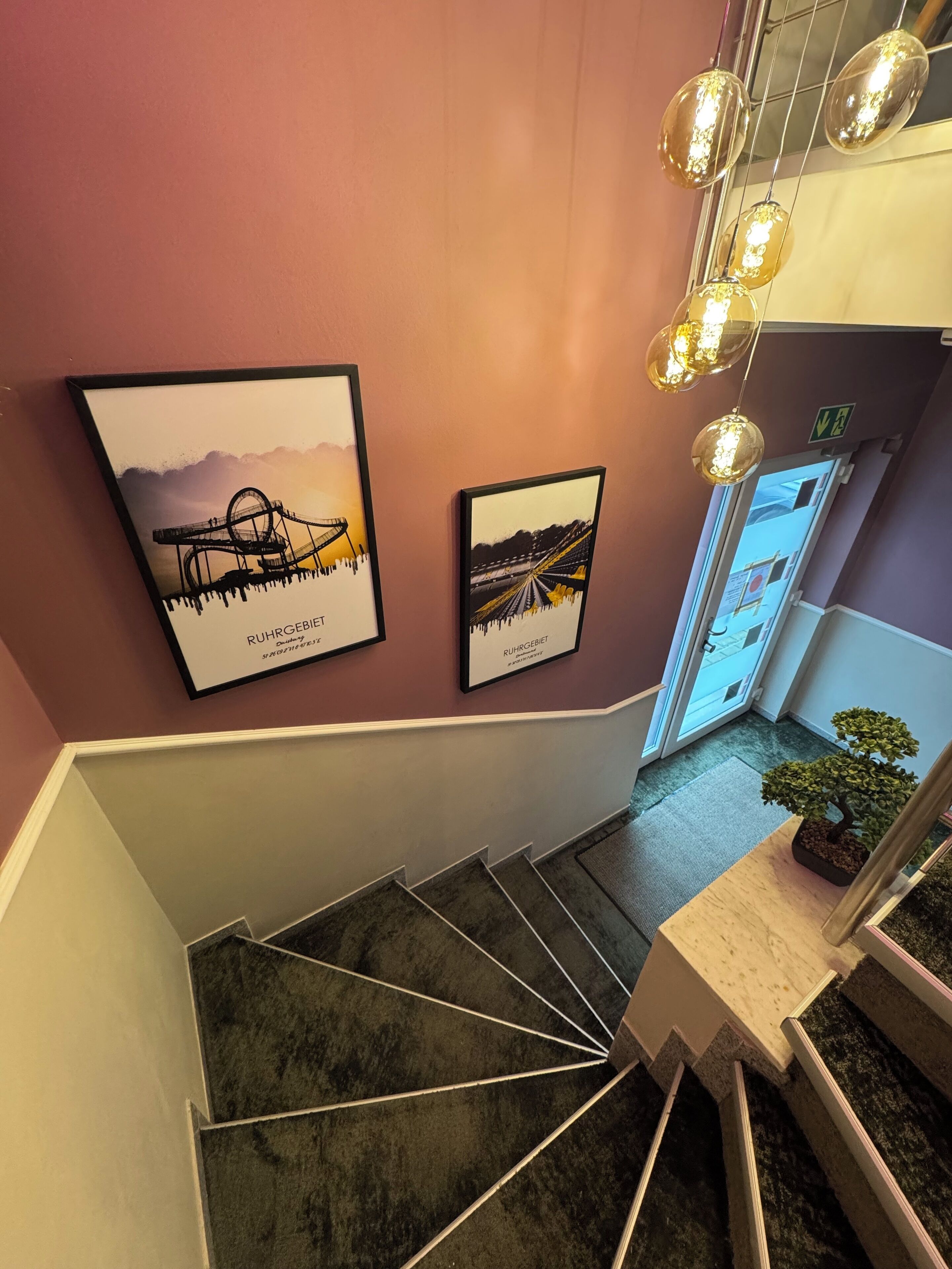 A staircase with a modern design, leading to a room with a view of a bridge. There are framed pictures on the wall, a small table with a plant, and hanging pendant lights.