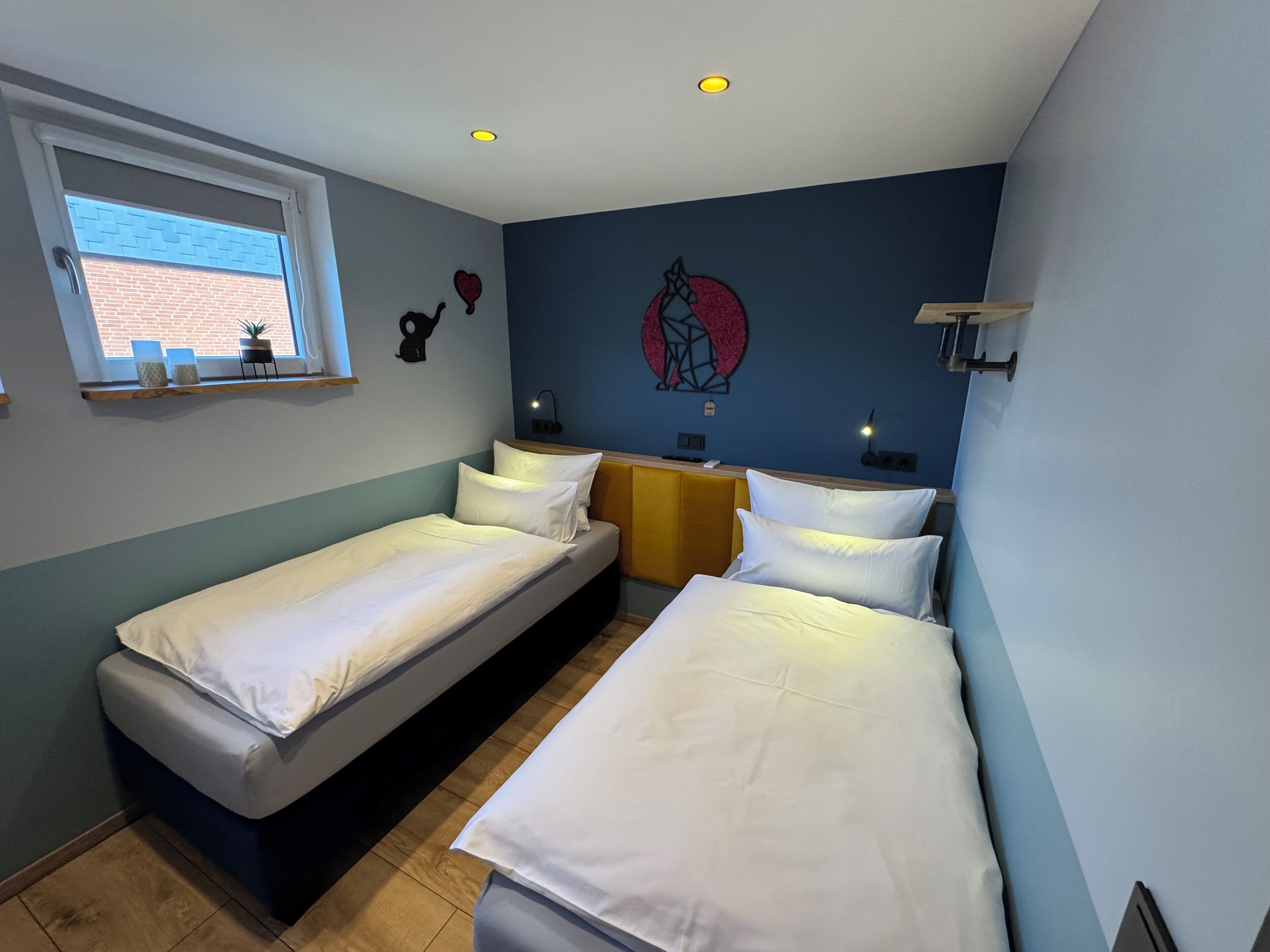 Ein Zimmer mit zwei Einzelbetten, einem Fenster mit Blick auf ein Ziegelgebäude und einer Wand mit geometrischem Muster.