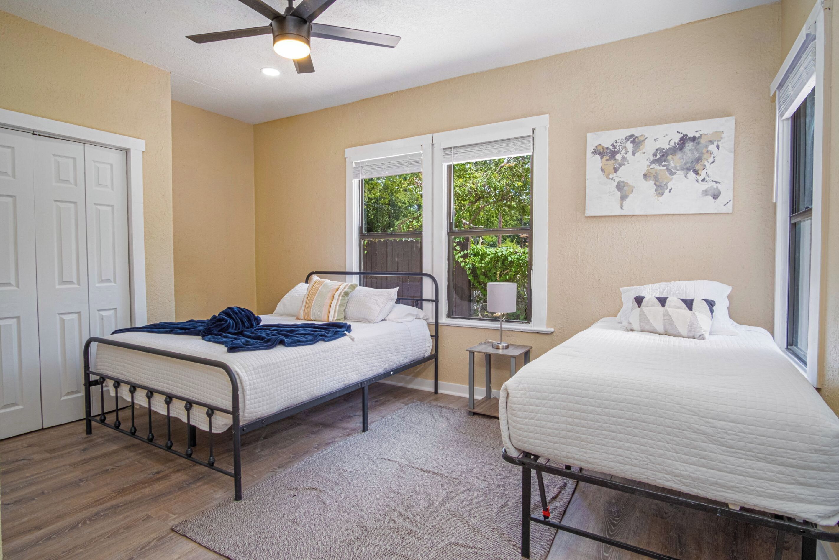 A bedroom with two beds, a ceiling fan, a world map on the wall, and a window with a view of greenery.