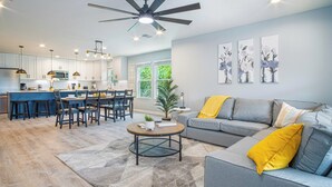 Una cocina moderna con armarios blancos, un comedor con mesa redonda y una sala de estar con sofá gris y almohadones amarillos.
