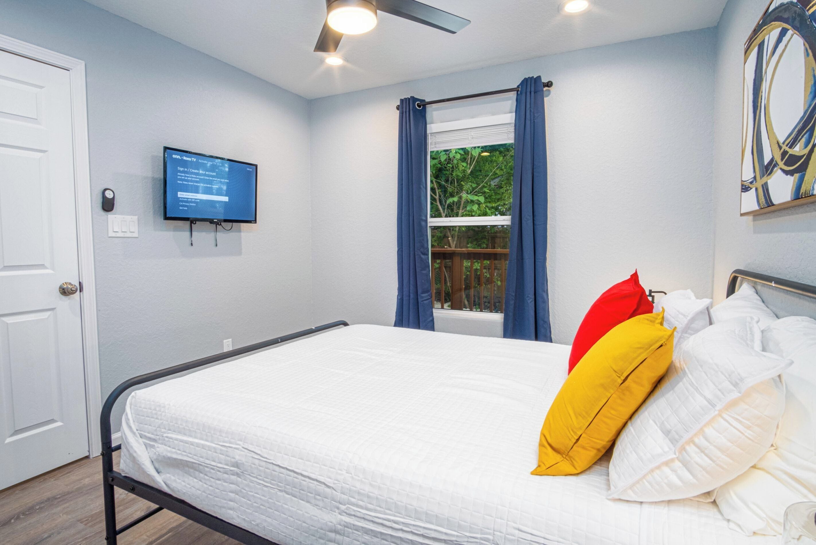 A bedroom with a bed, a ceiling fan, a window with curtains, a television on the wall, and a door.