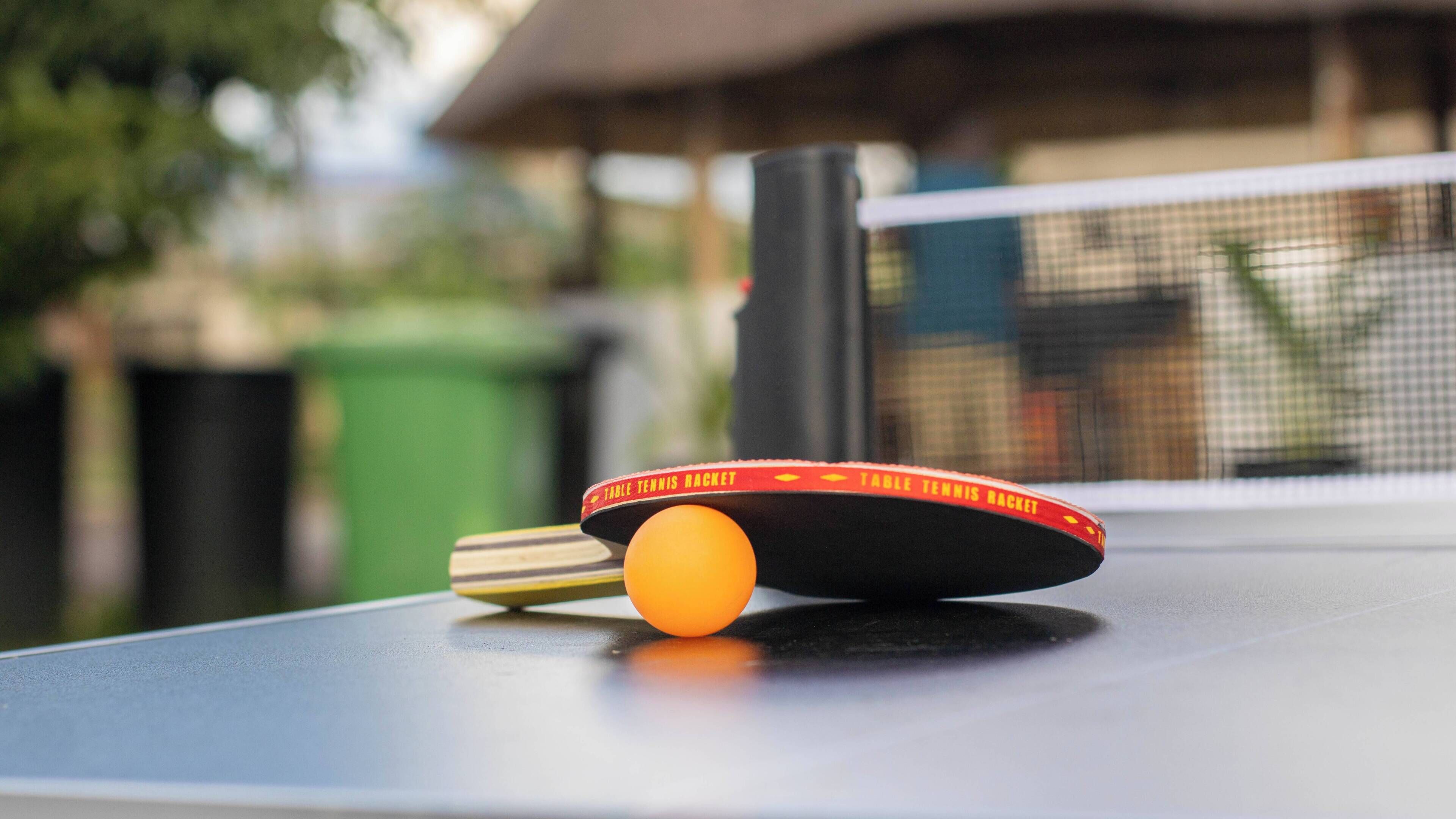 Ett bordtennisbord med ett racket och en boll, ett nät och en papperskorg i bakgrunden.