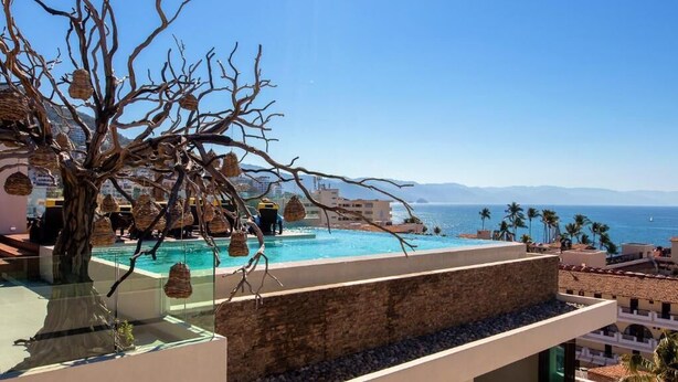 Une piscine sur le toit offrant une vue dégagée sur la mer et les montagnes, entourée d’immeubles et de palmiers.