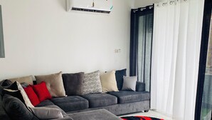 A modern living room with a grey sectional sofa, a ceiling fan, an air conditioning unit, and a large window with curtains. - Island vibes Oasis with a beautiful Pool (Kingston)