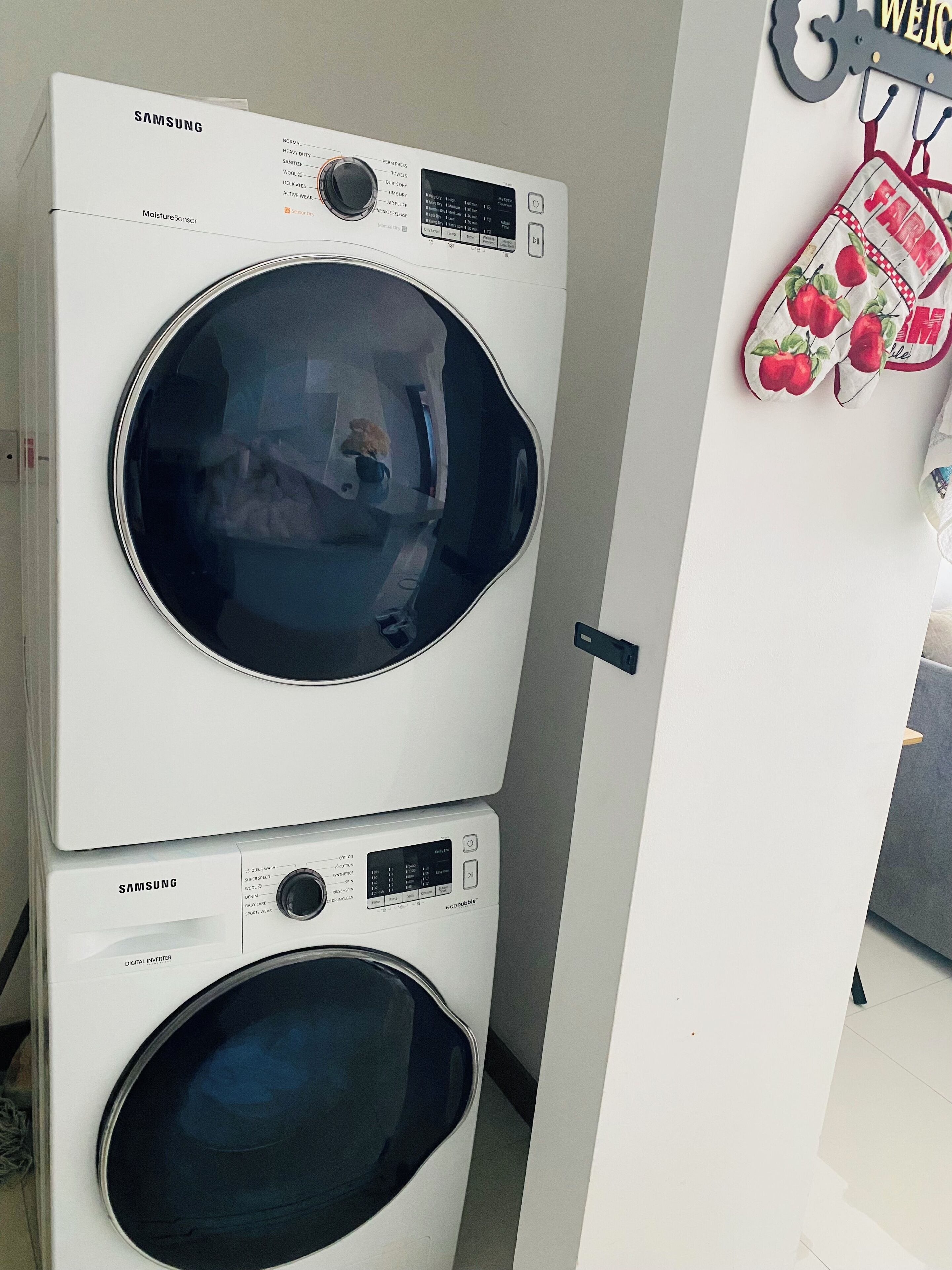Máquina de lavar e secar Samsung, ambas com abertura frontal, empilhadas numa lavanderia com um painel decorativo com tema de cozinha.