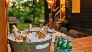 A metal bucket filled with ice and bottles of champagne and beer, placed on a wooden table outdoors with string lights in the background.