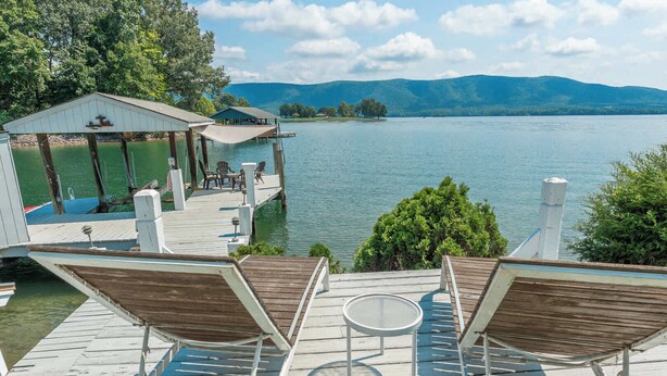 A wooden deck with two lounge chairs and a small table, overlooking a lake with a dock and a covered structure. - Stunning Lake Home w/ Private Dock, Spa, Fire Pit (Huddleston)