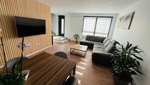 Un salon moderne avec un mur recouvert de panneaux de bois, un téléviseur à écran plat, un canapé gris, une table basse et un coin repas comprenant une table et des chaises en bois.