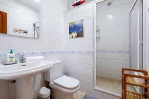 A bathroom with a sink, toilet, and shower. There is a painting on the wall and a wooden chair with towels. - Apartment California Ocean View (Santiago del Teide)