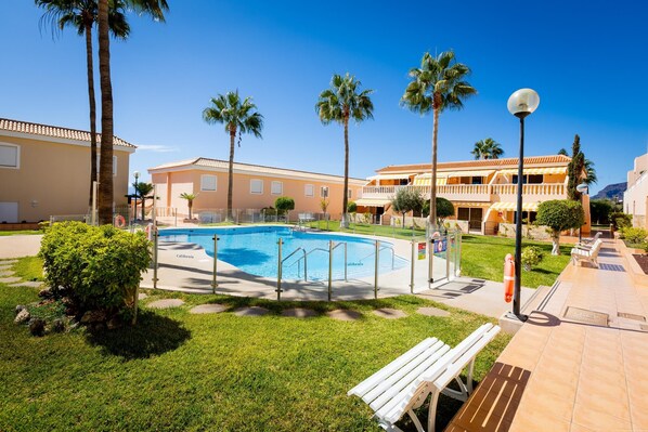 A swimming pool surrounded by palm trees and a building with a tiled roof. - Apartment California Ocean View (Santiago del Teide)