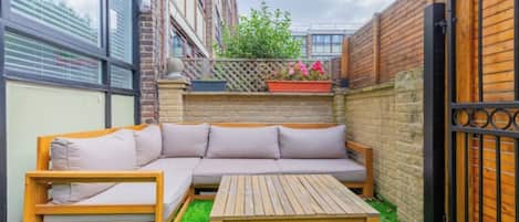 A small outdoor patio with a wooden bench, a wooden table, and a beige sectional sofa.