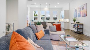 A spacious living room with a grey sofa, orange cushions, and a glass coffee table. A kitchen is visible in the background with modern appliances.