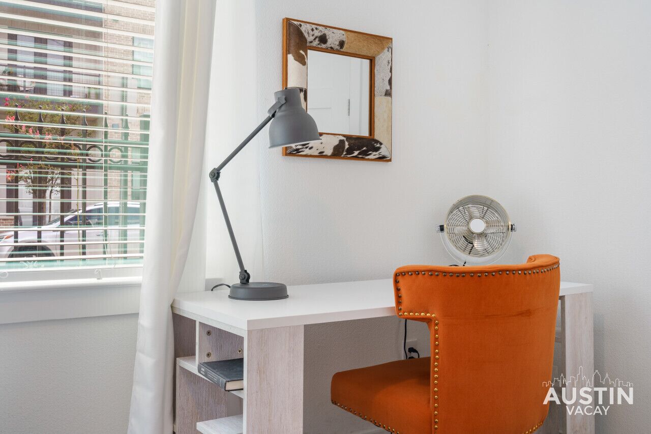 A white desk with a grey desk lamp, an orange chair, a wall-mounted mirror, and a standing fan.
