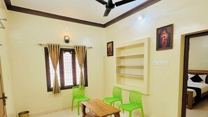 A room with a ceiling fan, a wooden table, green chairs, a window with curtains, and a bed in the adjacent room. - SSS Golden Resorts (Tarangambadi)