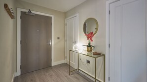 Un couloir doté d’un plancher en bois, une console en verre avec un cadre doré, un miroir et une plante en pot.