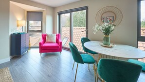 Un salon moderne avec un canapé rose, des fauteuils turquoise, une table en marbre et une grande fenêtre donnant sur un toit recouvert de tuiles.