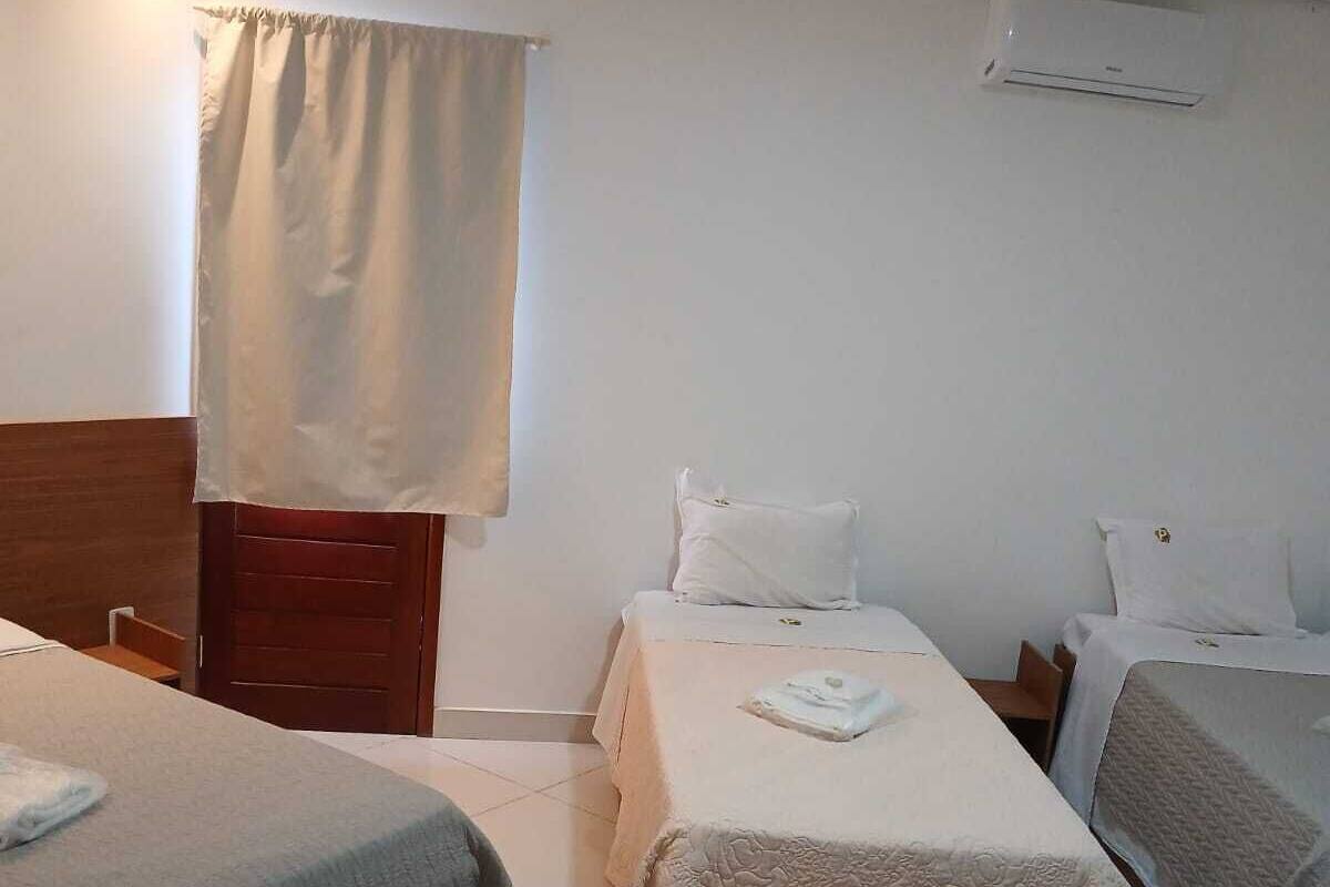 A hotel room with two beds, a wooden headboard, a window with curtains, a wall-mounted air conditioner, and a tiled floor.