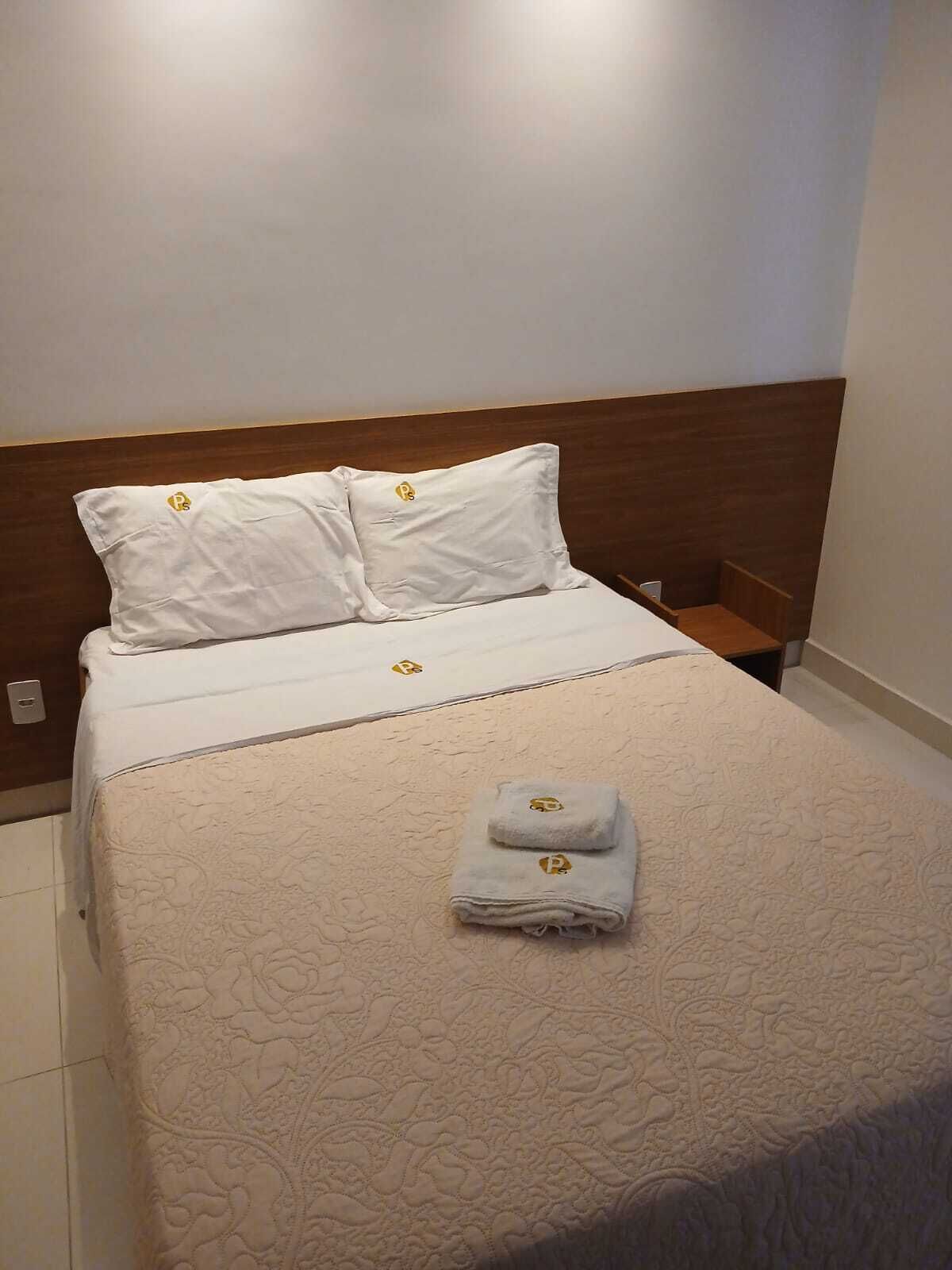 Een net opgemaakt bed met een beddekleed met patroon en twee kussens, een opgevouwen handdoek en een houten hoofdbord.