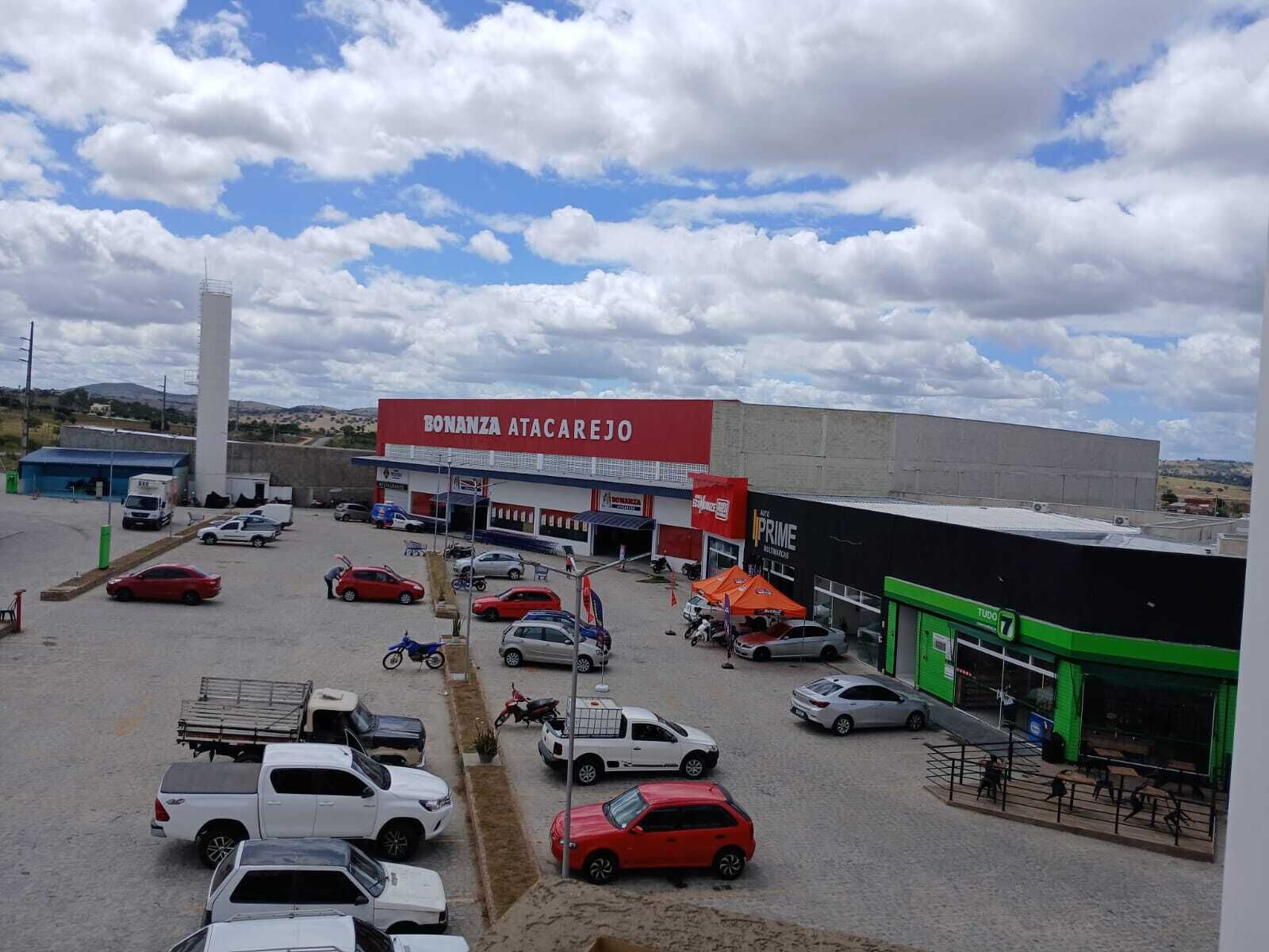 A parking lot with various vehicles, a large building with a sign reading "BONANZA ATACAREJO", and a green structure with a sign "TUDO".