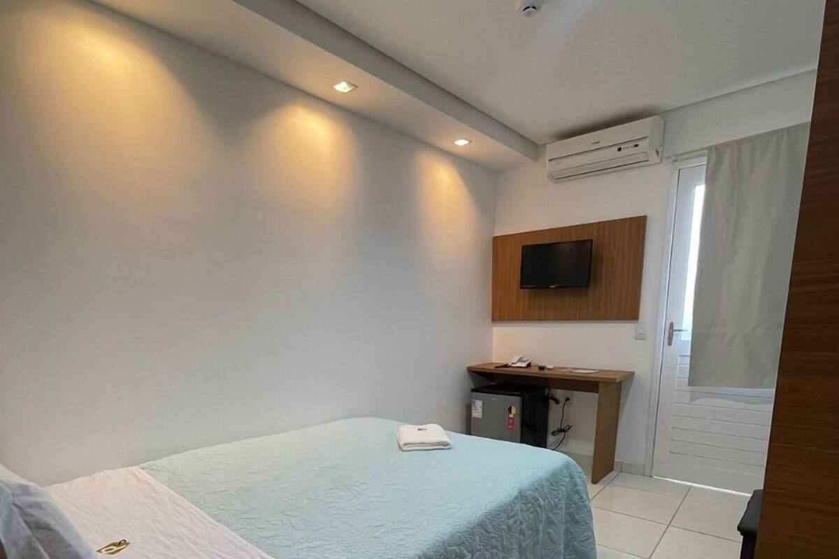 A hotel room with a bed, a desk, a television mounted on the wall, and an air conditioning unit.