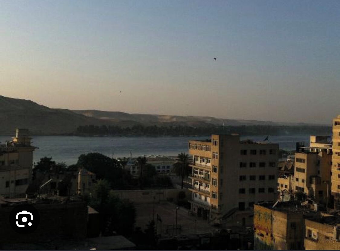 A coastal cityscape with buildings near the water, a clear sky, and a distant hill.