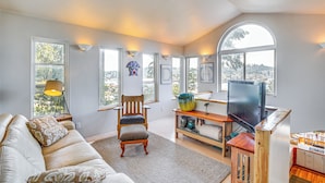 A living room with a sofa, armchair, TV, and large windows offering a view of trees and hills. - Gig Harbor Home: Large Deck, Outdoor Dining Area (Gig Harbor)