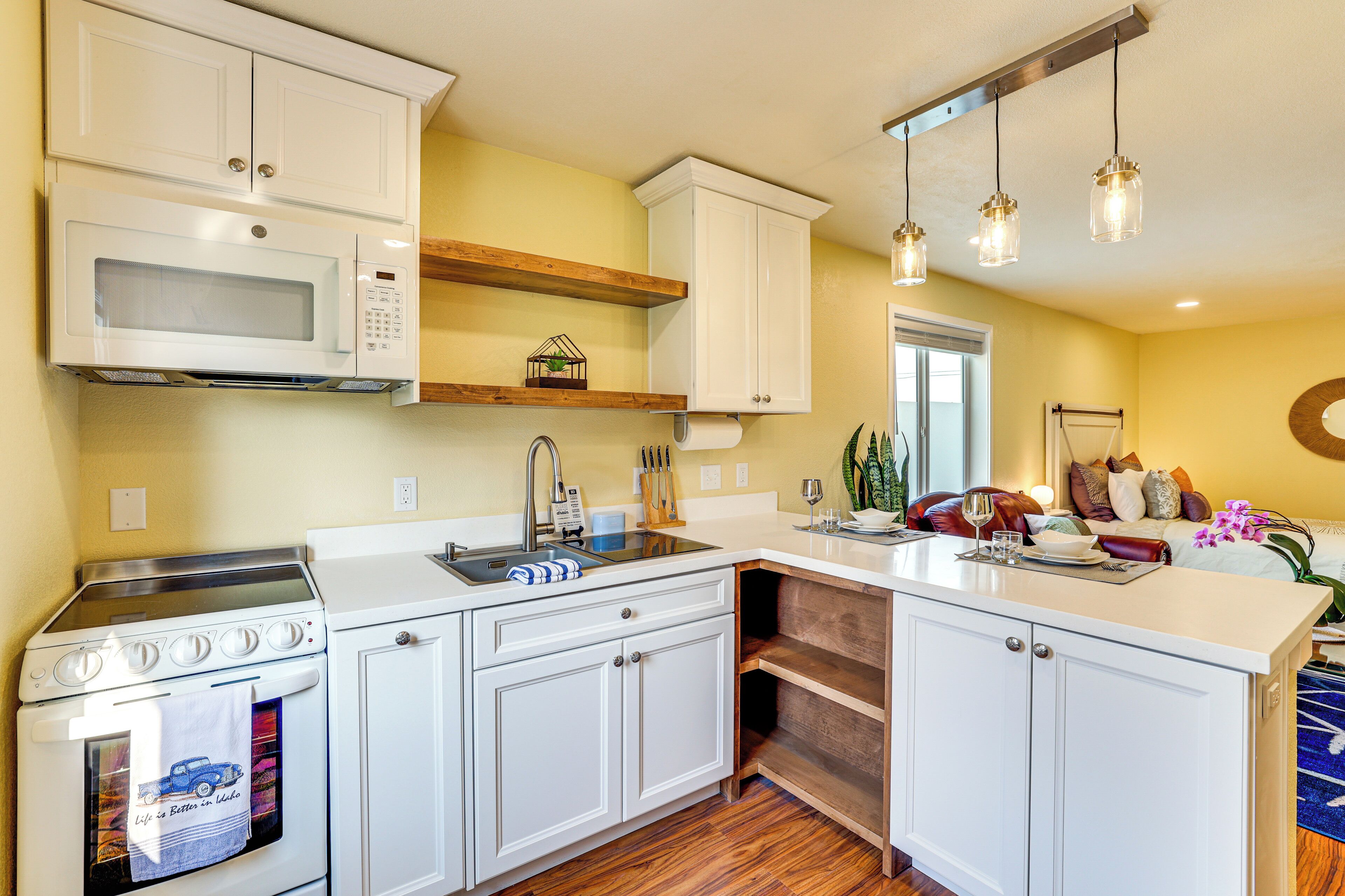 A modern kitchen with white cabinets, a microwave, a stove, and a sink. There is a wooden shelf above the sink and a hanging light fixture.