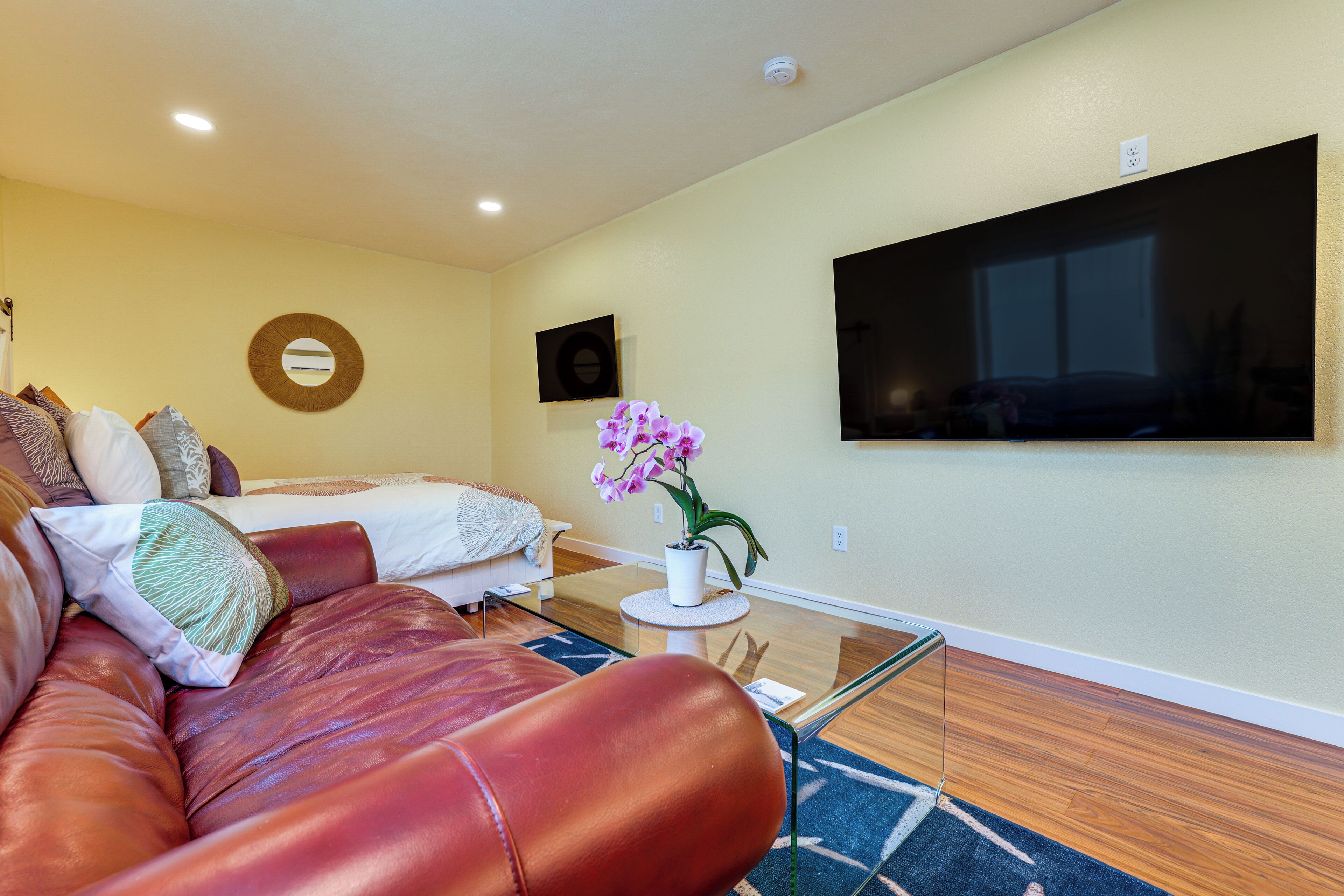 A modern living room with a leather sofa, a round mirror, a flat-screen TV, and a small table with a potted orchid.