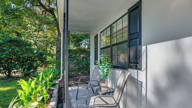 A covered porch with two chairs and a potted plant, surrounded by greenery. - Emerald Coast Home: Outdoor Entertainment Area! (Mary Esther)