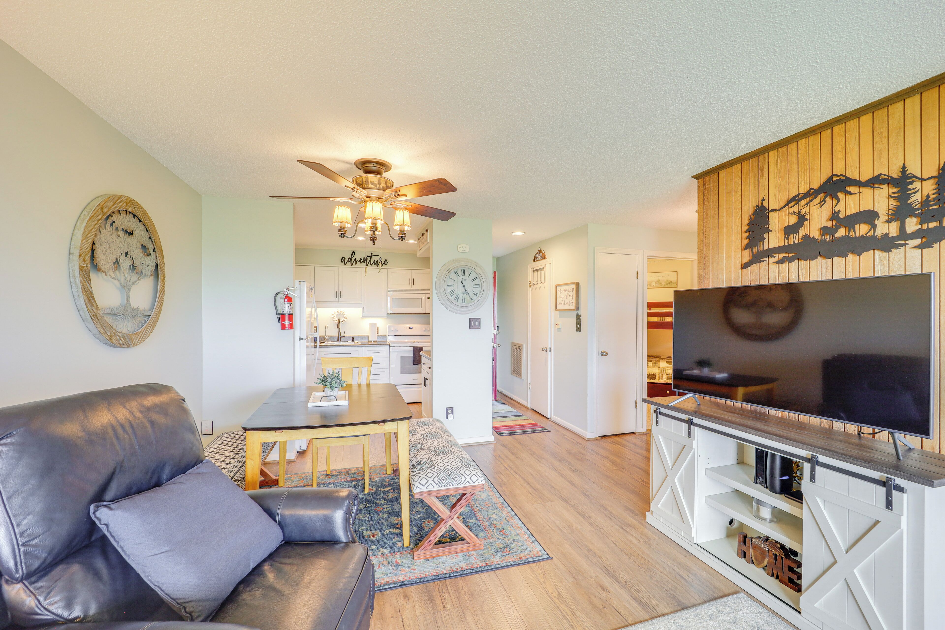 A modern living room with a leather sofa, a dining area with a table and chairs, a kitchen in the background, and a mounted TV on a wooden stand.