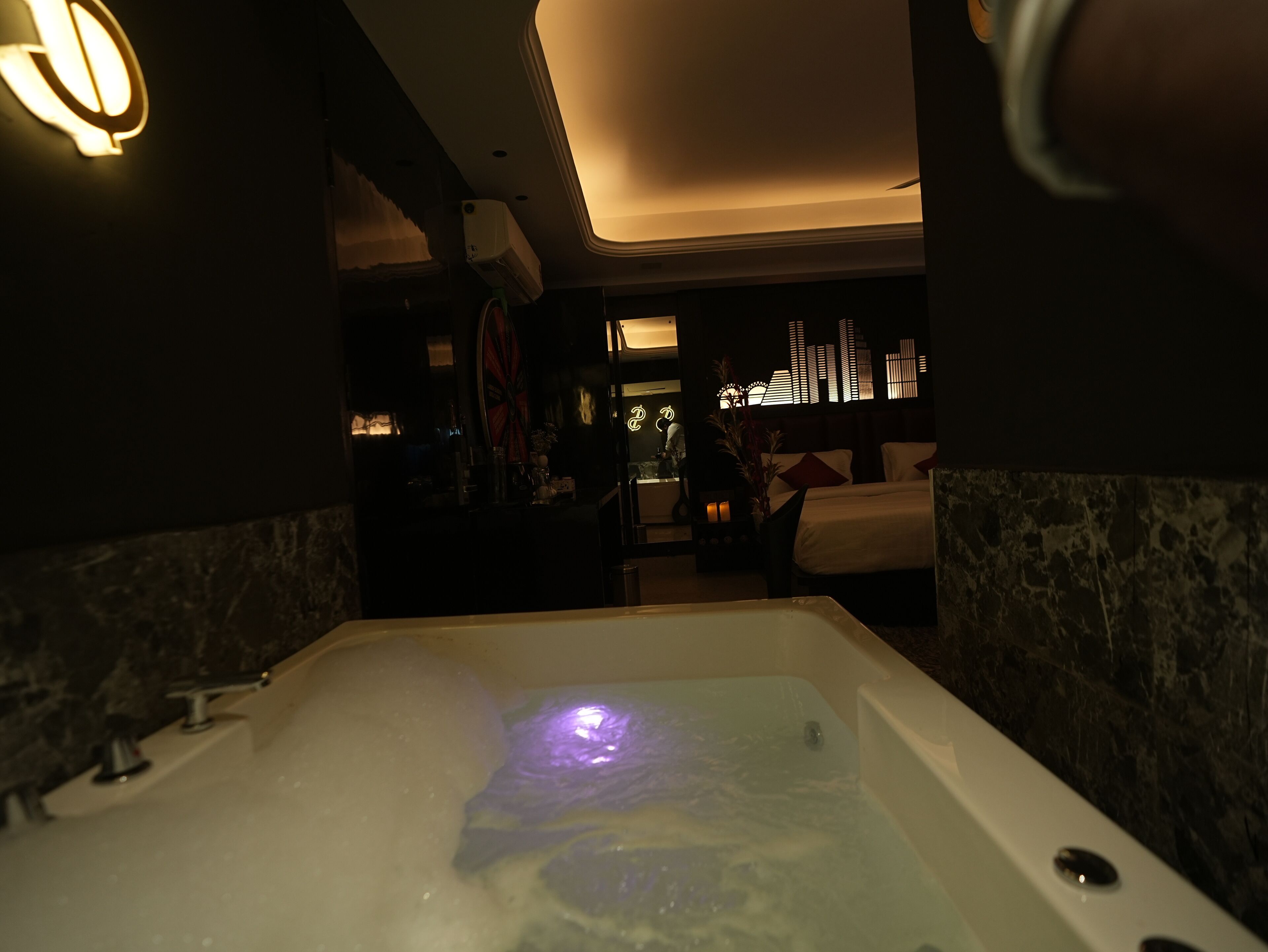 A dimly lit room with a large bathtub, a small seating area, and a decorative wall with a cityscape design.