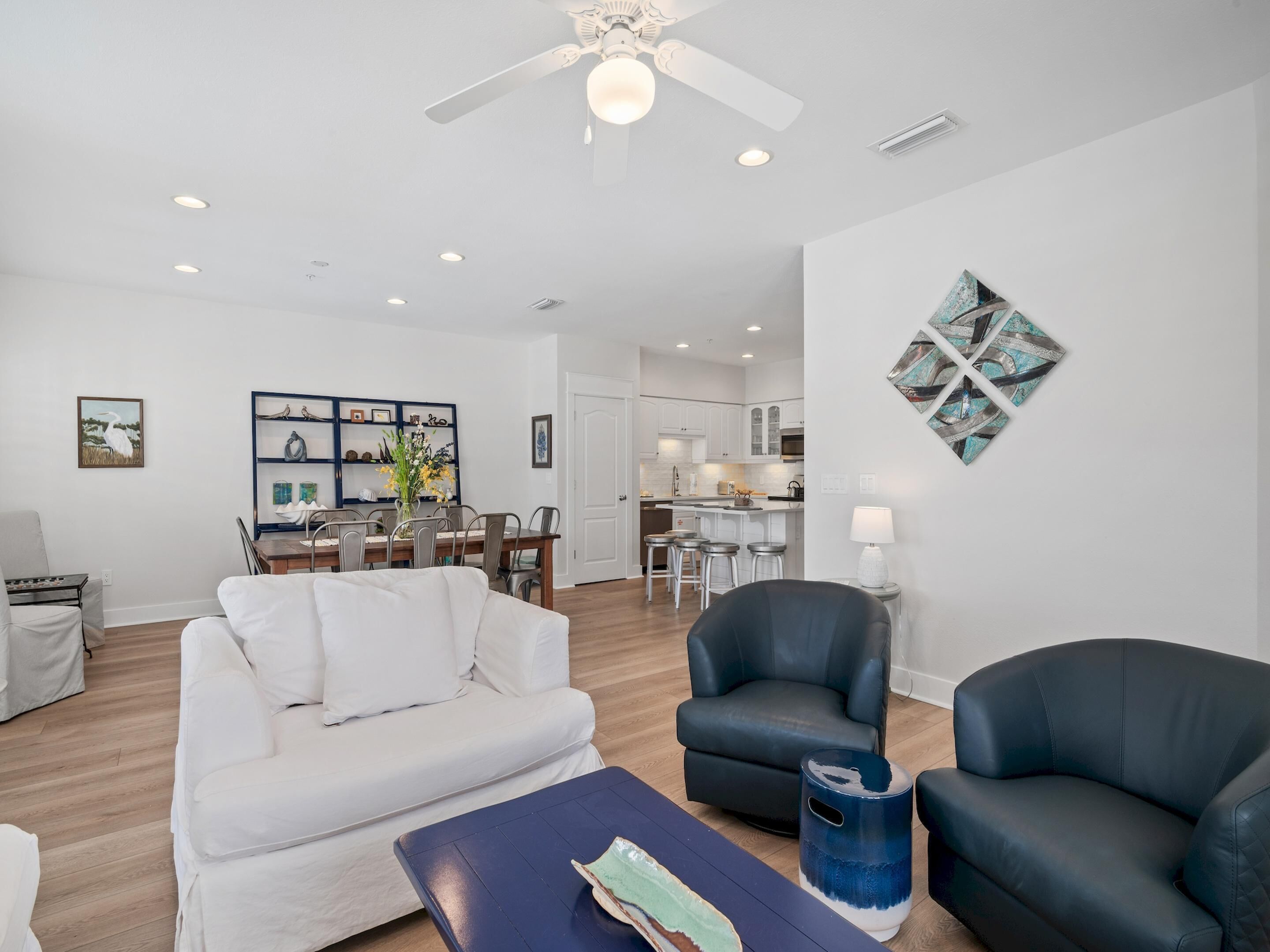 A spacious living room with a white sofa, a black leather armchair, and a coffee table. There is a dining area with a wooden table and chairs, and a kitchen in the background.