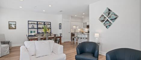 A spacious living room with a white sofa, a black leather armchair, and a coffee table. There is a dining area with a wooden table and chairs, and a kitchen in the background.