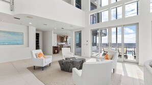 A spacious living area with white furniture, a large window overlooking a balcony with outdoor seating, and a kitchen in the background. - Sugar Sands Sanctuary: Breathtaking Beach Views, Private Pool, 6 Bedrooms ! (St. George Island)