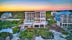 A beachfront property with multiple buildings, including a central white structure with large windows and a pool, surrounded by lush greenery and a clear sky. - Sugar Sands Sanctuary: Breathtaking Beach Views, Private Pool, 6 Bedrooms ! (St. George Island)