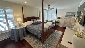 Una cama con dosel, un edredón estampado, una mesita de noche con lámpara, un aparador con espejo y una ventana con persianas.
