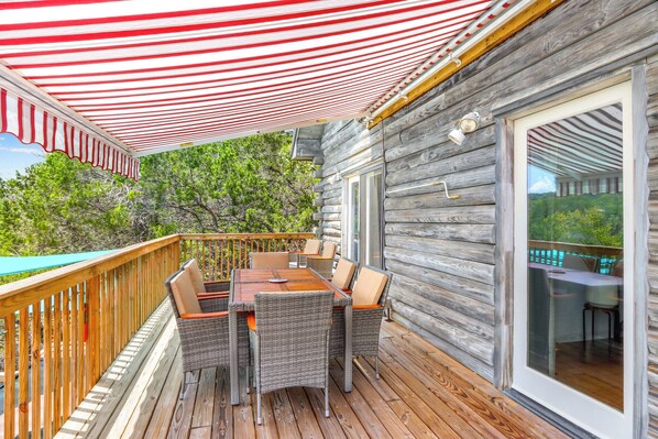 A wooden deck with a table and chairs, a glass door leading to a balcony, and a red and white striped awning. - Creekside Vista Escape - Lodge (Wimberley)