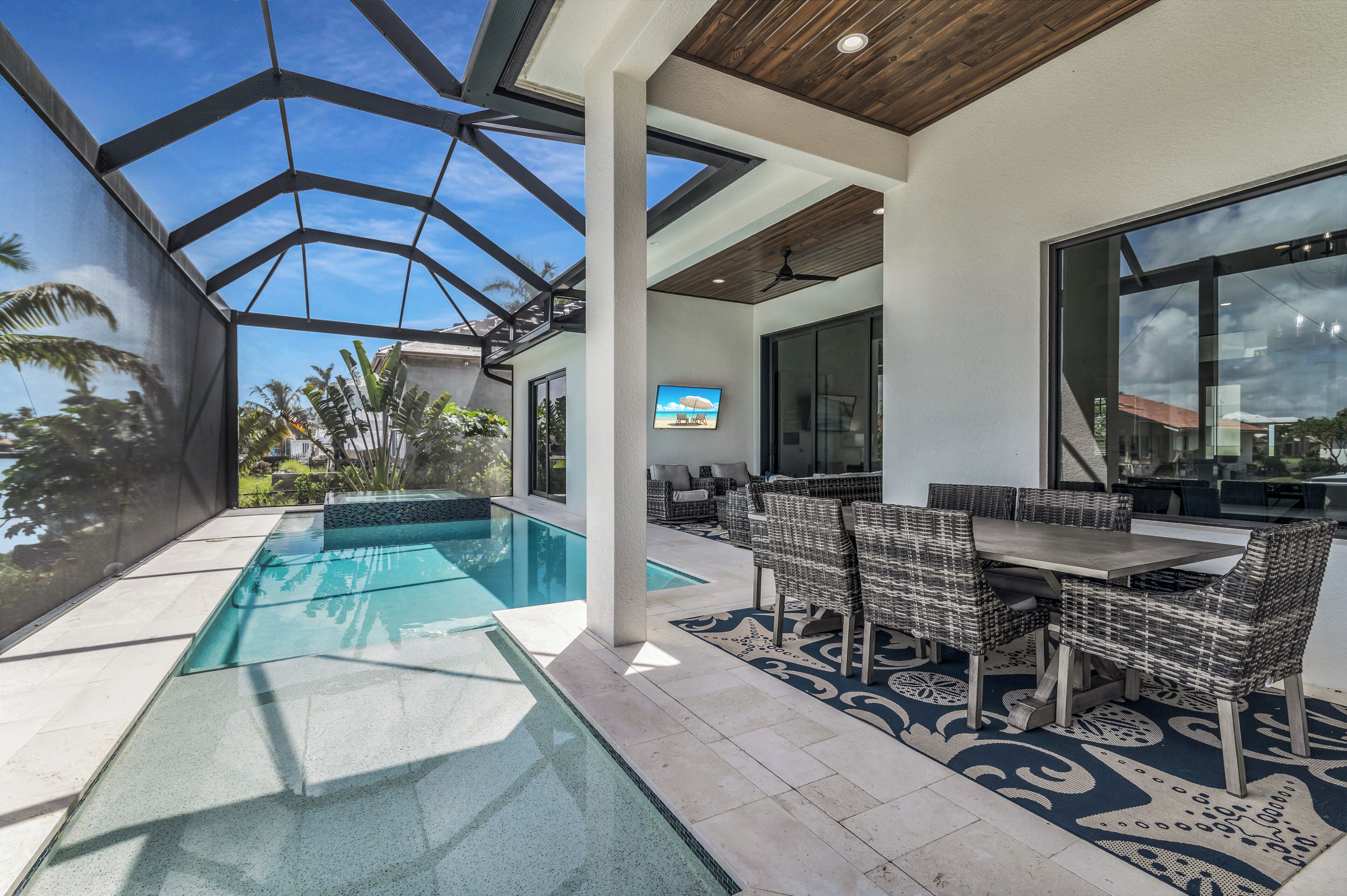 A modern outdoor pool area with a glass ceiling, a dining table with wicker chairs, and a view of the surrounding landscape.