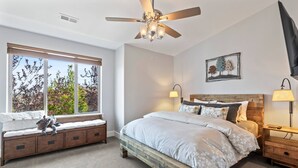 Un dormitorio con cama, un banco con cojines, ventilador de techo, mesita de noche, lámpara y ventana con vista a árboles.