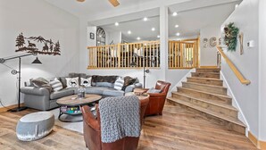Amplia sala de estar con un sofá gris, un sillón de cuero marrón y una mesa de centro de madera. Una escalera con barandilla de madera conduce a un nivel superior.