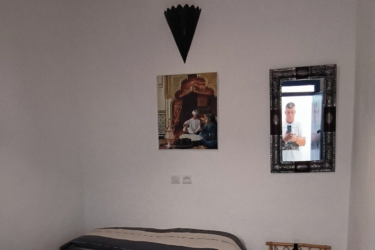 A bedroom with a bed, a hanging light fixture, a mirror, and a small wooden shelf.