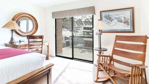 A hotel room with a bed, a wooden rocking chair, a desk with a lamp, a large window with a view of a snowy landscape, and a framed picture on the wall. - Ski In Ski Out 2BR Condo with Hot Tub and Pool (Park City)