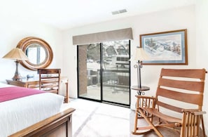Un cuarto de hotel con una cama, una mecedora de madera, un escritorio con una lámpara, un ventanal con vista a un paisaje nevado y un cuadro en la pared.