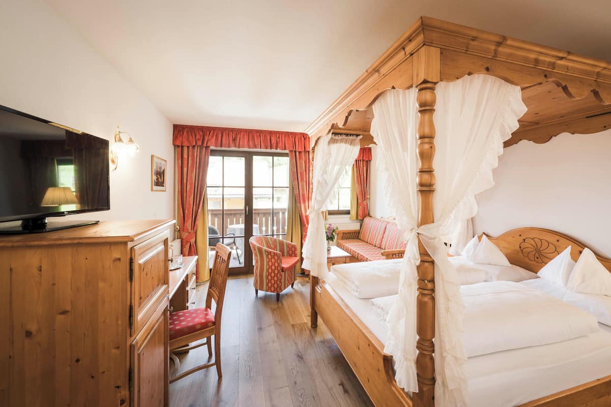Una cama con dosel y ropa de cama blanca, un escritorio de madera con un televisor, una silla roja y un sofá con cojines estampados.