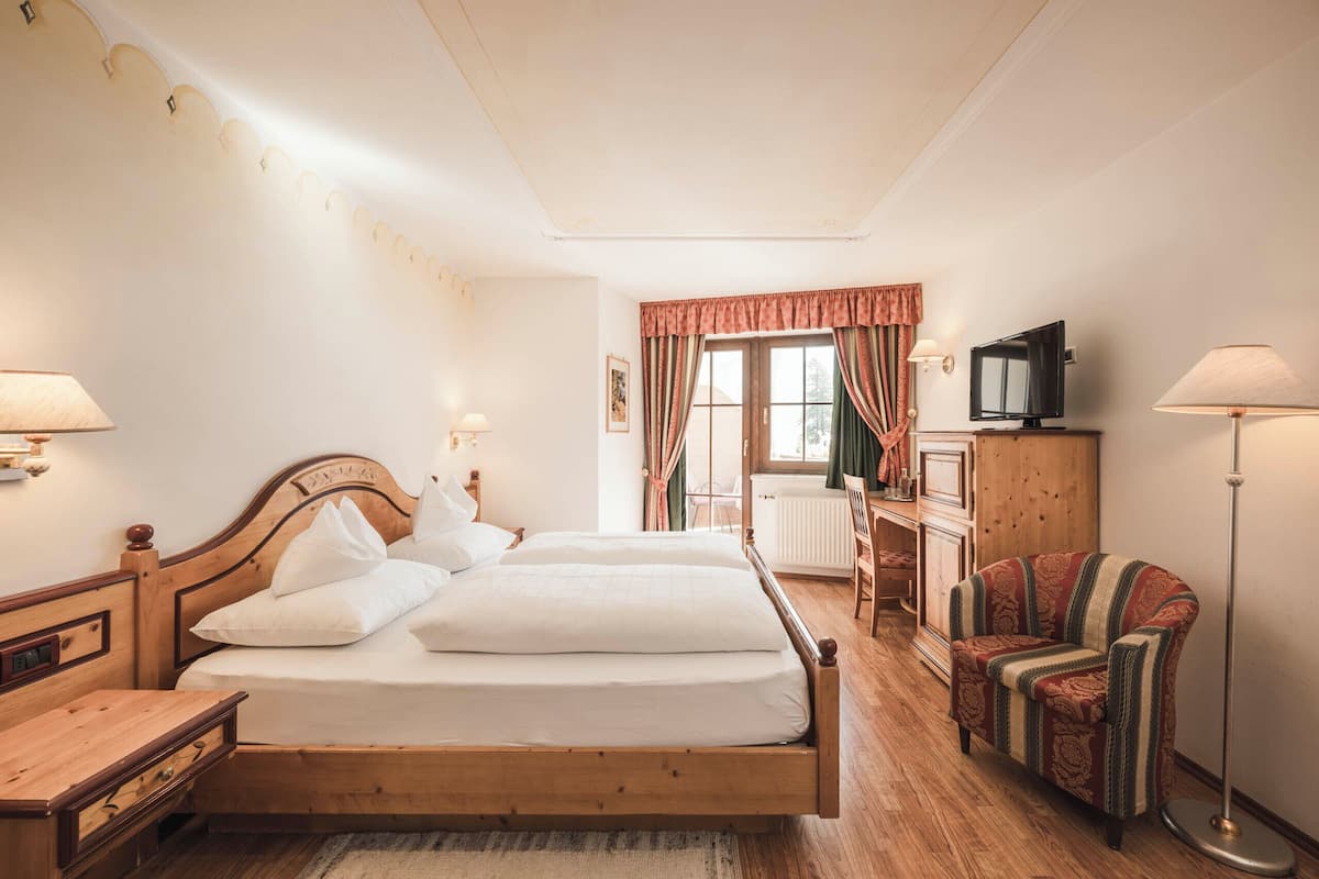 Una habitación de hotel con cama de madera, lámparas de noche, un televisor, una silla y un ventanal con cortinas.