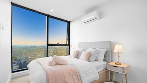 A modern bedroom with a large window, a bed with a headboard, a bedside table with a lamp, and a small storage basket.