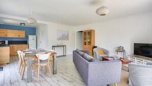 A modern living room with a grey sofa, dining table with chairs, and a kitchen area with wooden cabinets and a white refrigerator.