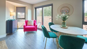 Un comedor moderno con una mesa redonda y sillas azul verdosas, un sillón rosa y un jarrón con flores.