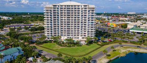 A high-rise hotel with multiple balconies, surrounded by a golf course, parking areas, and a body of water.